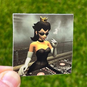 Pode incluir: Um adesivo com uma personagem de desenho animado em um vestido preto, óculos de sol e uma coroa de ouro, fumando um cigarro. A personagem está atrás de um equipamento de DJ. O fundo é uma imagem em tons de cinza de uma cidade.