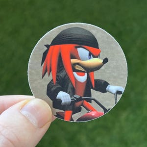 May include: A round sticker featuring a red and black cartoon character with a bandana, riding a motorbike. The character has a menacing expression and sharp teeth.