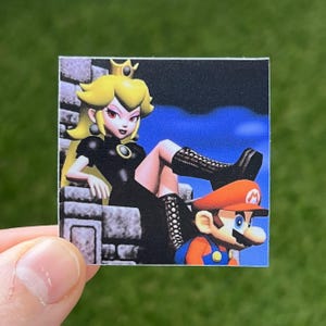 Op de afbeelding: Een sticker met een cartoon afbeelding van Prinses Peach in een zwarte jurk en zwarte laarzen, zittend op een troon met een kroon op haar hoofd. Mario zit voor haar op de grond, gekleed in zijn kenmerkende rode en blauwe outfit.