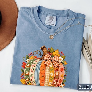 Puede incluir: Una camiseta de color azul vaquero con un gráfico de una calabaza decorativa con paneles verticales de varios patrones, incluyendo cuadros, flores y lunares, adornada con un lazo de cuadros, hojas de otoño, bayas y pequeñas setas.