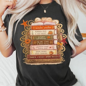 Peut inclure: Un t-shirt noir avec un motif représentant une pile de livres et des textes sur le thème de l'automne, notamment sweater weather, pumpkin spice & cozy nights, autumn adventures, falling leaves & books please, grateful heart et thankful blessed autumn obsessed.