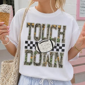 May include: A white short-sleeve t-shirt featuring the text TOUCH DOWN in a camouflage pattern. Between the words is a graphic of a football centered over a black and white checkered strip. The design is printed on the front of the shirt.