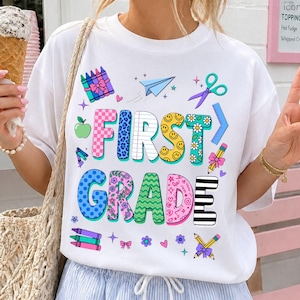 Peut inclure: Un t-shirt blanc à manches courtes avec une impression graphique colorée portant le texte FIRST GRADE. Le motif comprend diverses illustrations sur le thème de l'école telles que des crayons, un avion en papier, des ciseaux, une règle et un crayon.