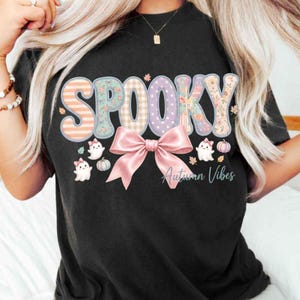 Peut inclure: Un t-shirt noir avec le mot SPOOKY écrit en grandes lettres pastel à motifs variés. Sous le texte se trouve un grand nœud rose, des petits fantômes blancs avec des nœuds roses, des citrouilles et des feuilles d'automne. Le texte Autumn Vibes est inscrit.