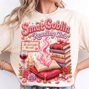 Op de afbeelding: Een crèmekleurig t-shirt met een afbeelding met de tekst "Smut Goblin Reading Club" en "We don't judge, We read". Het ontwerp bevat een stapel boeken, een wijnglas, een kaars, een roze kussen en rozen in roze en paarse tinten.