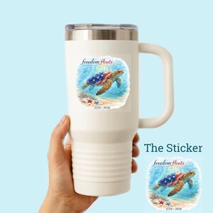 Op de afbeelding: Een witte reismok met een zilveren deksel en handvat. De mok heeft een sticker met een zeeschildpadontwerp, de schildpadschild is de Amerikaanse vlag. De sticker heeft ook de tekst "freedom floats" en "1776-2026".