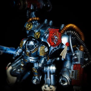 May include: A detailed, metallic miniature figure of a war machine, featuring silver, gold, and red accents. The figure has intricate armor, a shield with a red emblem, and a weapon. The overall design suggests a science fiction or fantasy theme.