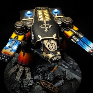 May include: A detailed miniature model of a war machine, featuring a black and gold color scheme with blue and yellow accents. The model has intricate details, including glowing blue lights and weapon details. The model is set on a rocky base.