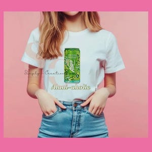 May include: A white t-shirt with a green and white Alani Nu Lime Flush energy drink can graphic. The text "Alani-aholic" is below the can. The model is wearing blue jeans.