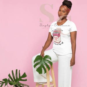 May include: White t-shirt with a graphic of a Red Bull pack, featuring the text "just a little something to take the edge off." The shirt is paired with white pants. The background is pink.