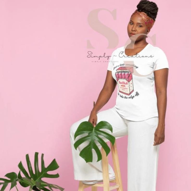 May include: White t-shirt with a graphic of a Red Bull pack, featuring the text "just a little something to take the edge off." The shirt is paired with white pants. The background is pink.