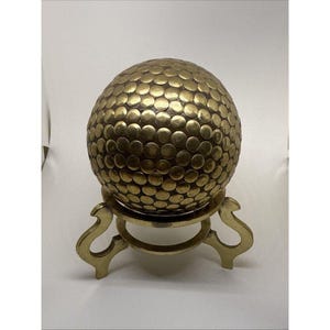 Peut inclure: Un objet sphérique en laiton avec une surface texturée composée de nombreuses petites indentations circulaires, ressemblant à une balle de golf. La sphère repose sur un support décoratif en laiton à trois pieds avec des pieds courbés.