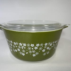 Puede incluir: Una fuente de horno redonda de vidrio verde oliva con dos asas laterales integradas y una tapa de vidrio transparente. El exterior de la fuente presenta un patrón floral blanco repetitivo que consiste en pequeñas flores de cinco pétalos y hojas.