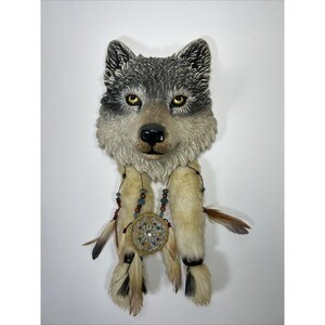 Könnte beinhalten: Eine Wandskulptur in Form eines Wolfskopfes mit grauer und weißer Fellstruktur, gelben Augen und schwarzer Nase. Darunter hängen zwei beige, fellbedeckte Anhängsel, verziert mit braunen und blauen Perlen, Federn und einem runden Traumfänger-Element.
