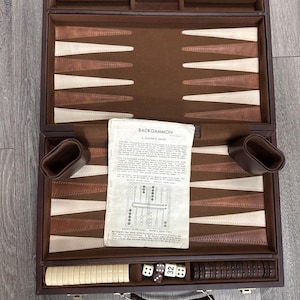 May include: A brown leather-effect backgammon set in an open carrying case featuring a brown and cream triangular point pattern. The set includes a set of cream and dark brown checkers, four dice, a doubling cube, and a paper instruction guide titled BACKGAMMON A PLAYER'S GUIDE.