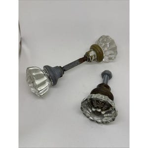 May include: Two clear glass door knobs with faceted edges and metallic bases. One knob is attached to a long, thin, rusted metal spindle, while the other is a single knob featuring a short, threaded metal bolt with a hexagonal nut attached to the end.