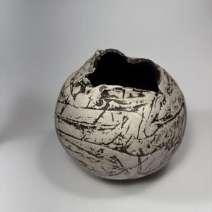 May include: A spherical ceramic vessel with a textured, off-white exterior featuring deep, dark brown linear markings and cracks. The opening at the top is irregular and jagged, revealing a dark, hollow interior space within the rounded form.