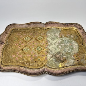 May include: A rectangular tray with a scalloped edge, featuring a distressed pink border and a central gold-toned surface. The centre displays a repeating diamond pattern with small green and pink floral accents, partially overlaid with a faded white design.