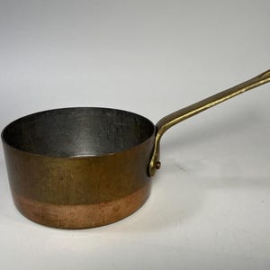 May include: A round copper saucepan with a dark, weathered patina on the exterior and a metallic interior. The pan features a long, angled brass handle securely attached to the side with a single visible rivet, extending outward from the body.