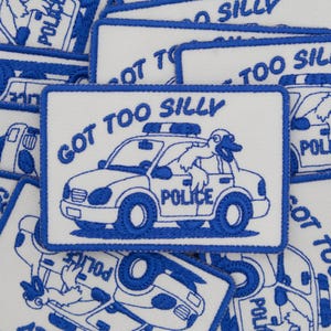 Könnte beinhalten: Ein rechteckiger gestickter Aufnäher mit weißem Hintergrund und blauem Rand. Das Motiv zeigt eine Comic-Gans, die aus dem Fenster eines Polizeiautos lehnt, mit dem Text GOT TOO SILLY darüber und dem Wort POLICE auf der Autotür.