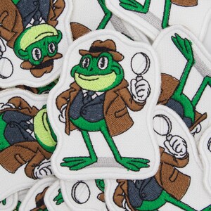 Könnte beinhalten: Eine Sammlung bestickter Stoffaufnäher mit einem grünen Frosch als Detektiv. Der Frosch trägt einen braunen Fedora und Trenchcoat über einer dunklen Weste mit Krawatte und hält eine weiße Lupe. Das Motiv befindet sich auf weißem Grund.