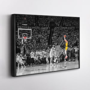 May include: A framed black and white photograph of a basketball game in action. A player in a yellow uniform is mid-air, shooting a basketball. The crowd is in the background, and the image is in a black frame.