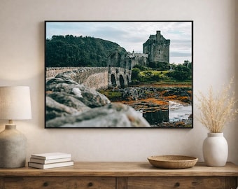 May include: A framed wall art print depicting Eilean Donan Castle in Scotland. The image features a stone bridge leading to the historic fortress, surrounded by lush green hills and a calm body of water with rocky foreground details under a cloudy sky.