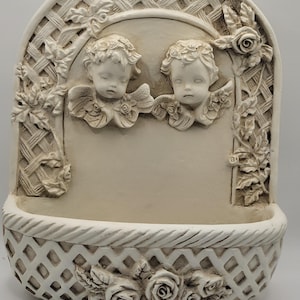 May include: Off-white wall-mounted decorative piece featuring two cherubic figures with wings. The design includes a lattice arch, floral accents, and a basket-like base with rose details. A vintage-style home decor item.