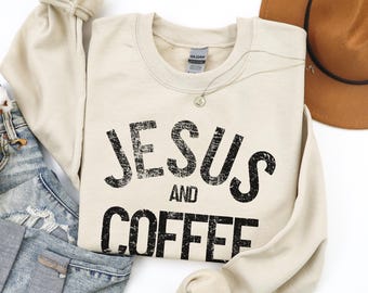May include: A cream-coloured sweatshirt featuring the text "JESUS AND COFFEE" in distressed black lettering. The sweatshirt is laid on blue denim jeans and a brown hat.
