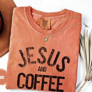 May include: A coral-colored t-shirt with the words "JESUS AND COFFEE" printed in black. A gold necklace with a pendant rests on the shirt. A brown hat and dried palm leaves are in the background.