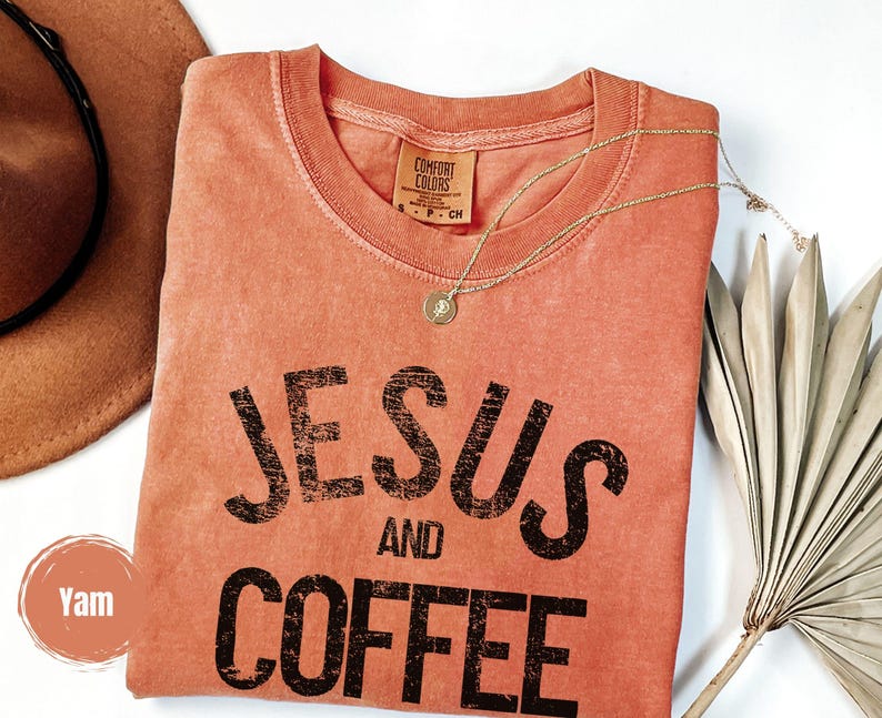 May include: A coral-colored t-shirt with the words "JESUS AND COFFEE" printed in black. A gold necklace with a pendant rests on the shirt. A brown hat and dried palm leaves are in the background.