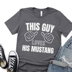 May include: A dark gray t-shirt with white text that reads "This Guy Loves His Mustang". The text is accompanied by two white hand gestures, one on each side of the text.