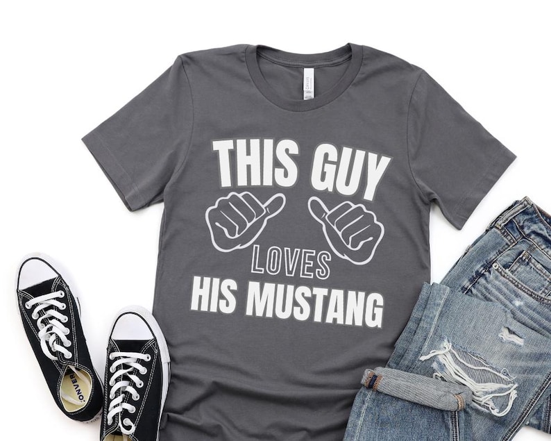 May include: A dark gray t-shirt with white text that reads "This Guy Loves His Mustang". The text is accompanied by two white hand gestures, one on each side of the text.