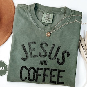May include: A moss green t-shirt with the words "JESUS AND COFFEE" printed in black. A gold necklace with a pendant rests on the shirt. A brown hat and dried leaves are in the background.