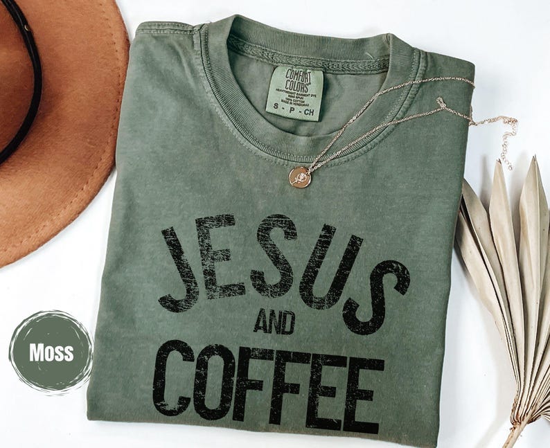 May include: A moss green t-shirt with the words "JESUS AND COFFEE" printed in black. A gold necklace with a pendant rests on the shirt. A brown hat and dried leaves are in the background.