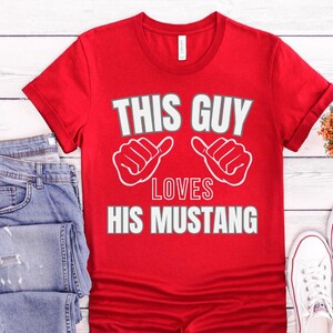 May include: Red t-shirt with white text that says "This Guy Loves His Mustang". The text is surrounded by two white thumbs up.