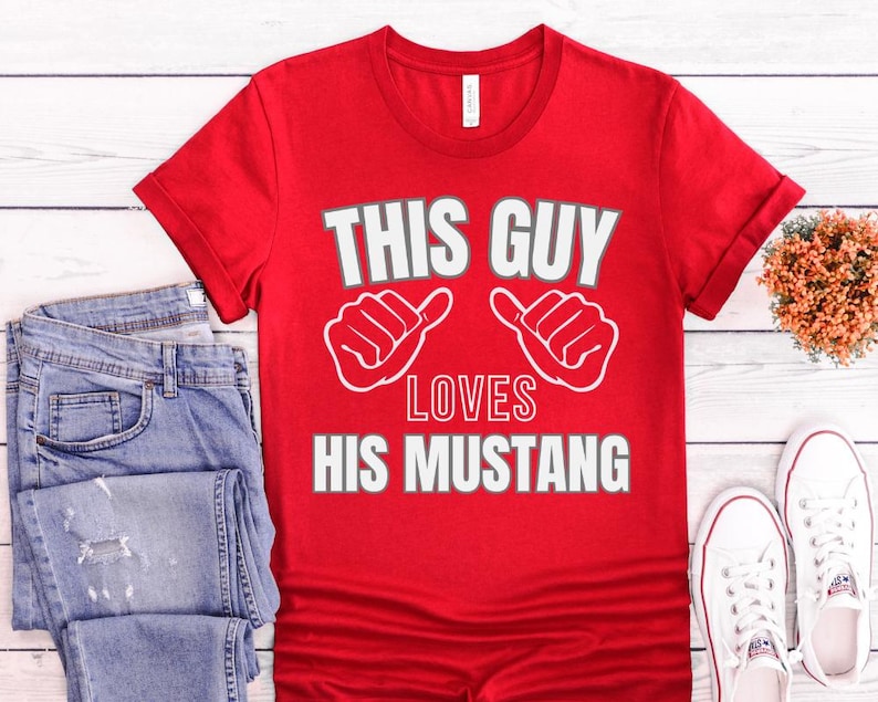 May include: Red t-shirt with white text that says "This Guy Loves His Mustang". The text is surrounded by two white thumbs up.