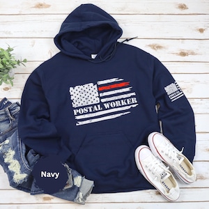 Puede incluir: Sudadera con capucha azul marino de "Postal Worker" con un diseño de bandera estadounidense desgastada y el texto "POSTAL WORKER" en blanco. Cuenta con una franja roja y un pequeño gráfico de bandera en la manga. Incluye bolsillo delantero y capucha con cordón.