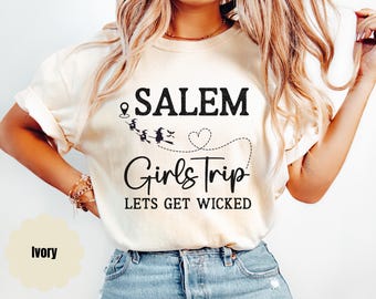 May include: Ivory-colored t-shirt with the text "Salem Girls Trip Lets Get Wicked" in black. The design includes a location pin, witch silhouettes on broomsticks, and a heart. The shirt is made of a soft material.