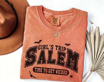 May include: A coral-colored t-shirt with the text "GIRL'S TRIP SALEM TIME TO GET WICKED" in black lettering. The shirt features bat and spider graphics. A gold necklace and a brown hat are also visible.