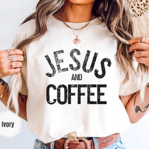 May include: Ivory-colored t-shirt with the text "JESUS AND COFFEE" in distressed black lettering. The shirt is a neutral color and has a classic crew neck design. The shirt is a casual wear item.