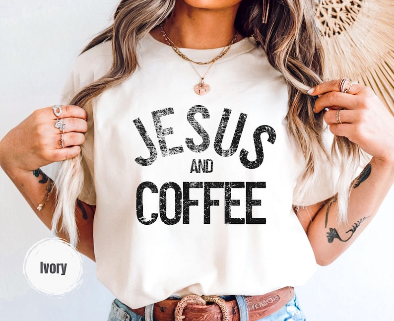 May include: Ivory-colored t-shirt with the text "JESUS AND COFFEE" in distressed black lettering. The shirt is a neutral color and has a classic crew neck design. The shirt is a casual wear item.