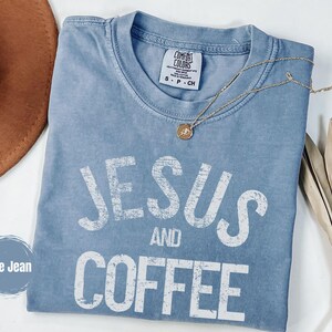 May include: A blue jean Comfort Colors t-shirt with the words "JESUS AND COFFEE" in white, distressed lettering. A gold necklace with a pendant is draped across the shirt. A brown hat and dried plant are in the background.