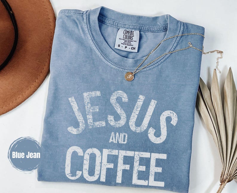 May include: A blue jean Comfort Colors t-shirt with the words "JESUS AND COFFEE" in white, distressed lettering. A gold necklace with a pendant is draped across the shirt. A brown hat and dried plant are in the background.