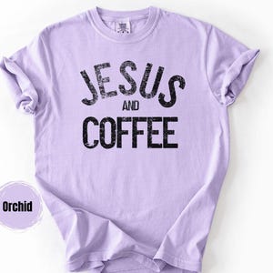 May include: Orchid-colored t-shirt with the words "JESUS AND COFFEE" printed in distressed black lettering. The shirt has short sleeves and a crew neck. The shirt is made of a soft material.