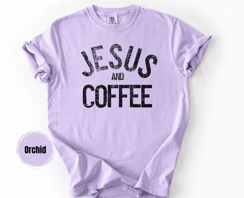 May include: Orchid-colored t-shirt with the words "JESUS AND COFFEE" printed in distressed black lettering. The shirt has short sleeves and a crew neck. The shirt is made of a soft material.