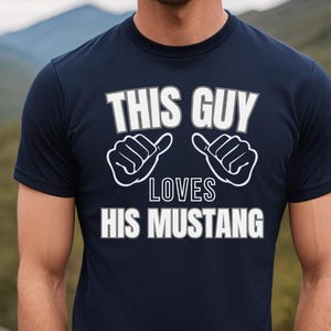 May include: A navy blue t-shirt with white text that reads "This Guy Loves His Mustang". The text is surrounded by two white thumbs up symbols.