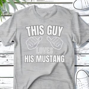 May include: Gray t-shirt with the text "This Guy Loves His Mustang" in white. The text is surrounded by two thumbs up symbols.