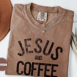 May include: Espresso-colored t-shirt with the words "JESUS AND COFFEE" in black block letters. A silver necklace with a pendant rests on the shirt. The shirt has a Comfort Colors tag. A brown hat and dried leaves are in the background.