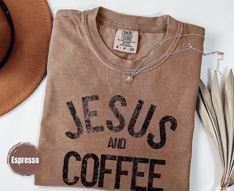 May include: Espresso-colored t-shirt with the words "JESUS AND COFFEE" in black block letters. A silver necklace with a pendant rests on the shirt. The shirt has a Comfort Colors tag. A brown hat and dried leaves are in the background.